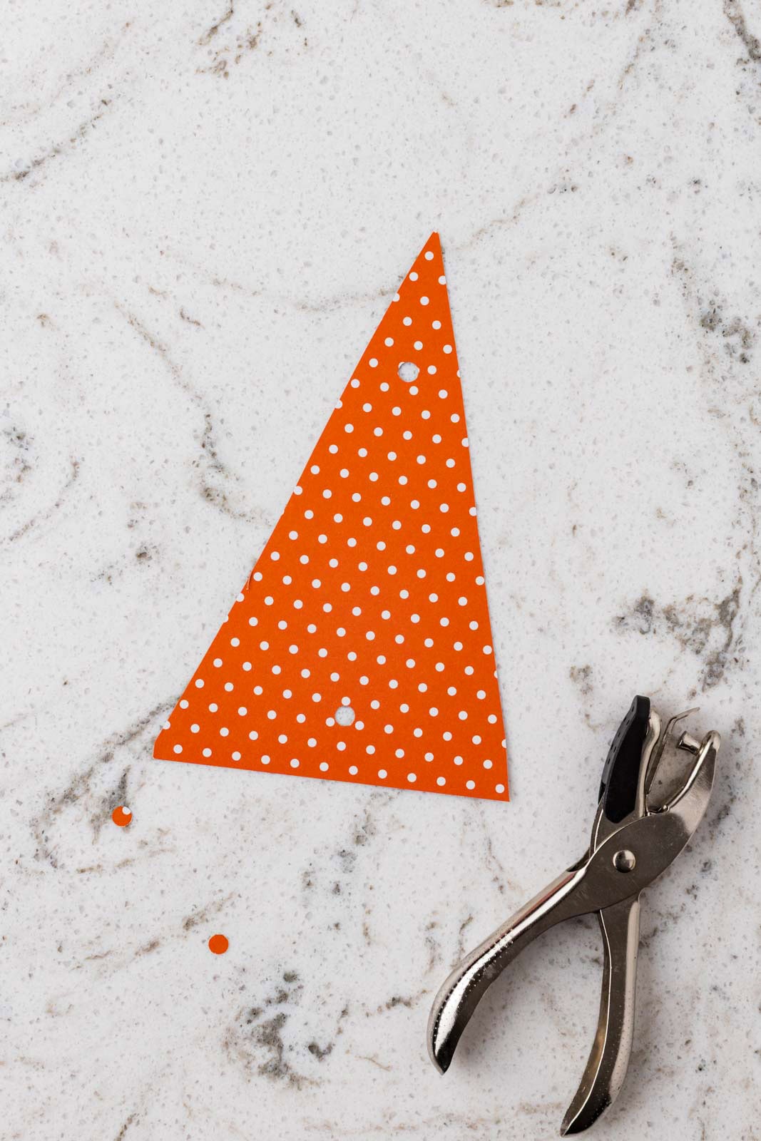 Orange polka dot paper triangle with punched holes next to a metal hole punch on a marble surface.