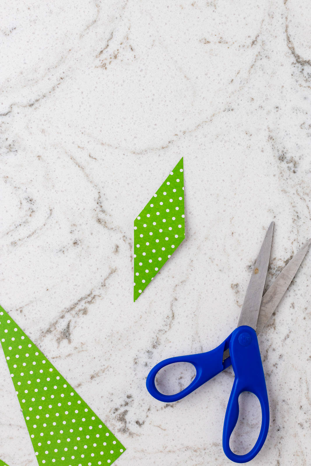Green polka dot paper flag cut into shape beside blue-handled scissors and paper scraps on a marble countertop.