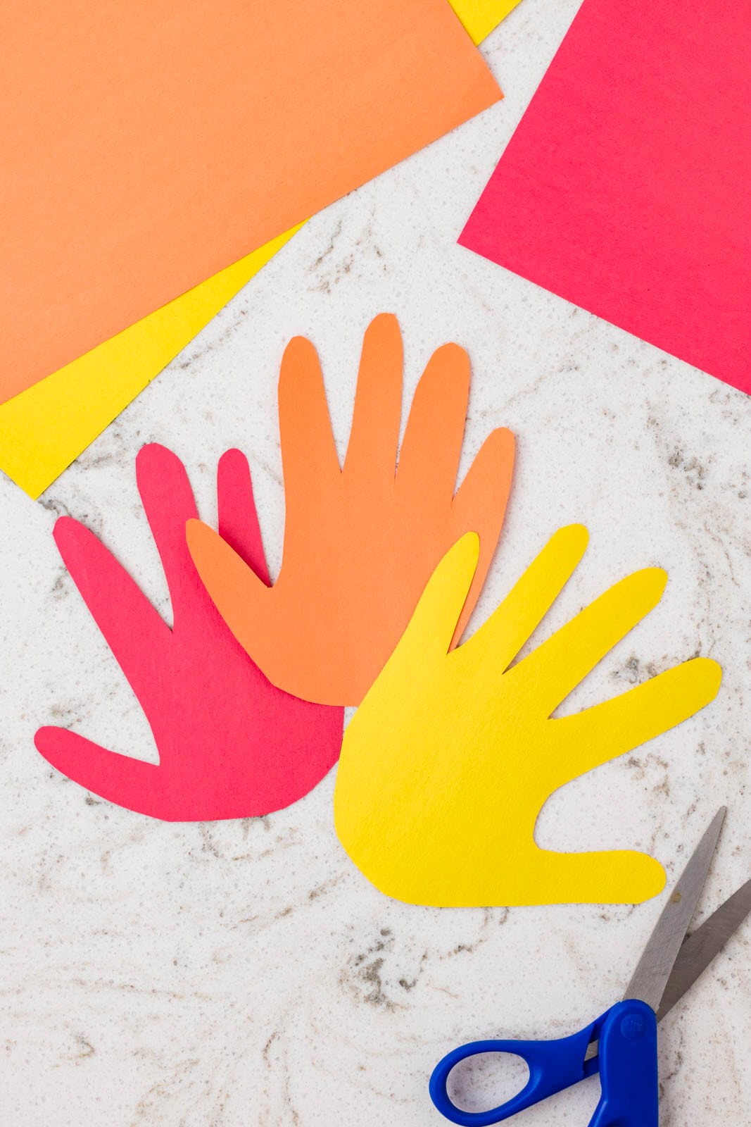 Colorful handprint cutouts in red, orange, and yellow arranged on a table with craft paper and scissors.