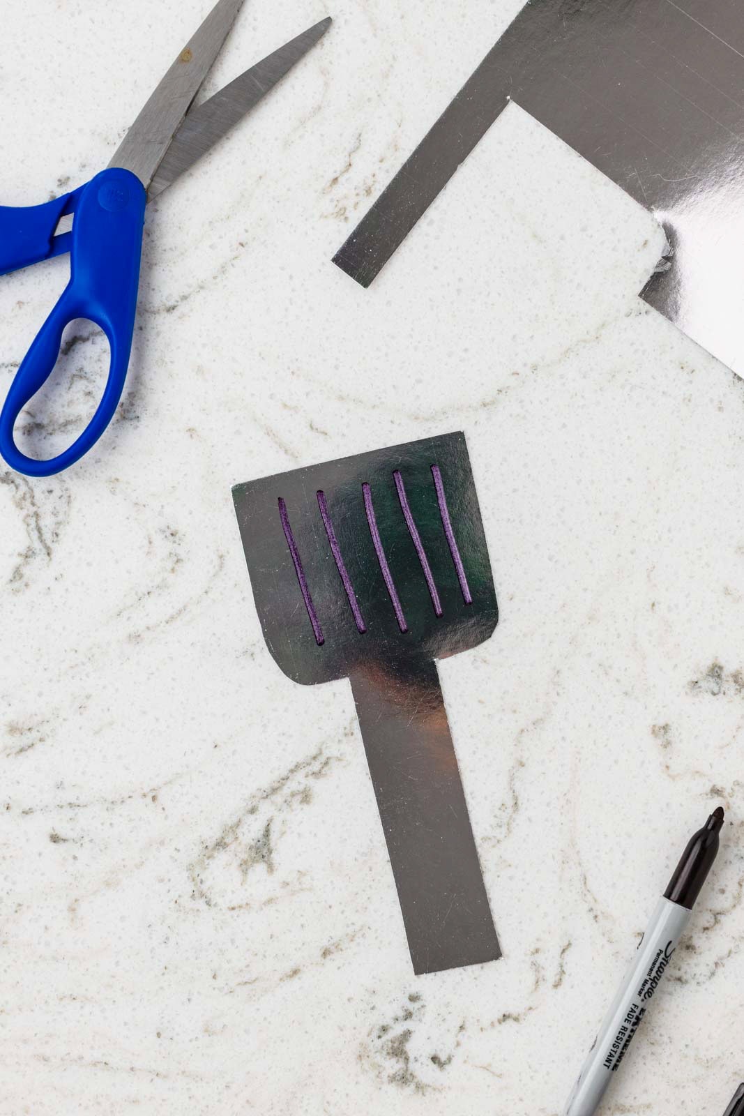 Metallic paper cut into a spatula shape with drawn grill lines, alongside scissors and a marker.