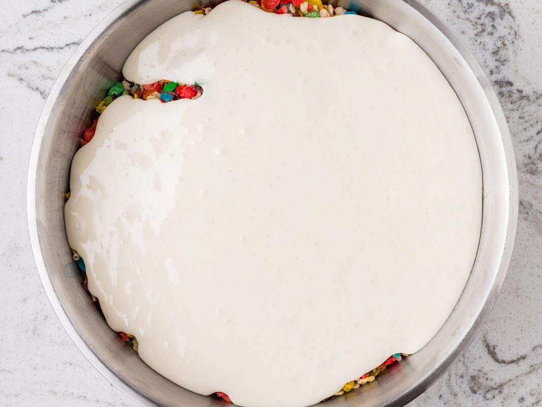 Melted marshmallow mixture being poured over colorful cereal and candy pieces in a mixing bowl for rainbow Rice Krispies treats.
