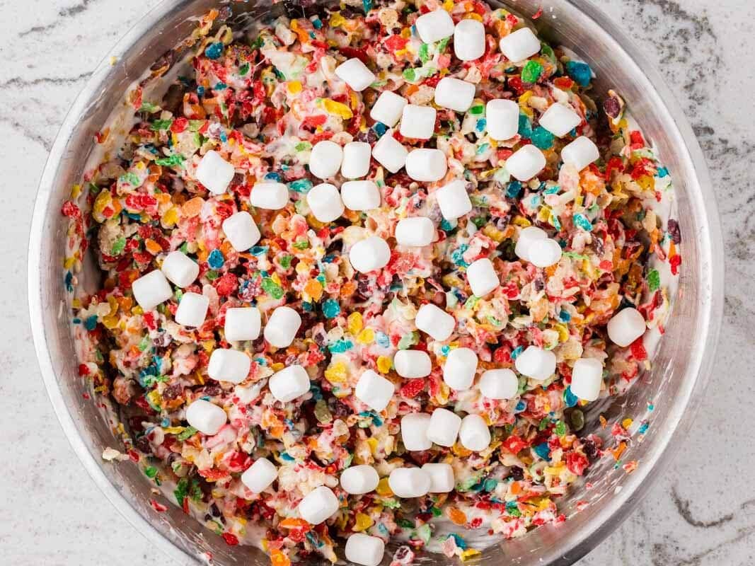 Large bowl of rainbow Rice Krispies treat mixture with fruity cereal, mini marshmallows, and chocolate candies coated in melted marshmallow.