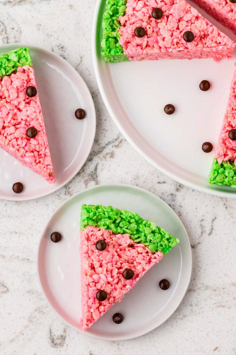 Watermelon-shaped Rice Krispies treat slice on a plate, featuring a pink center, bright green rind, and chocolate candy pieces arranged as watermelon seeds.