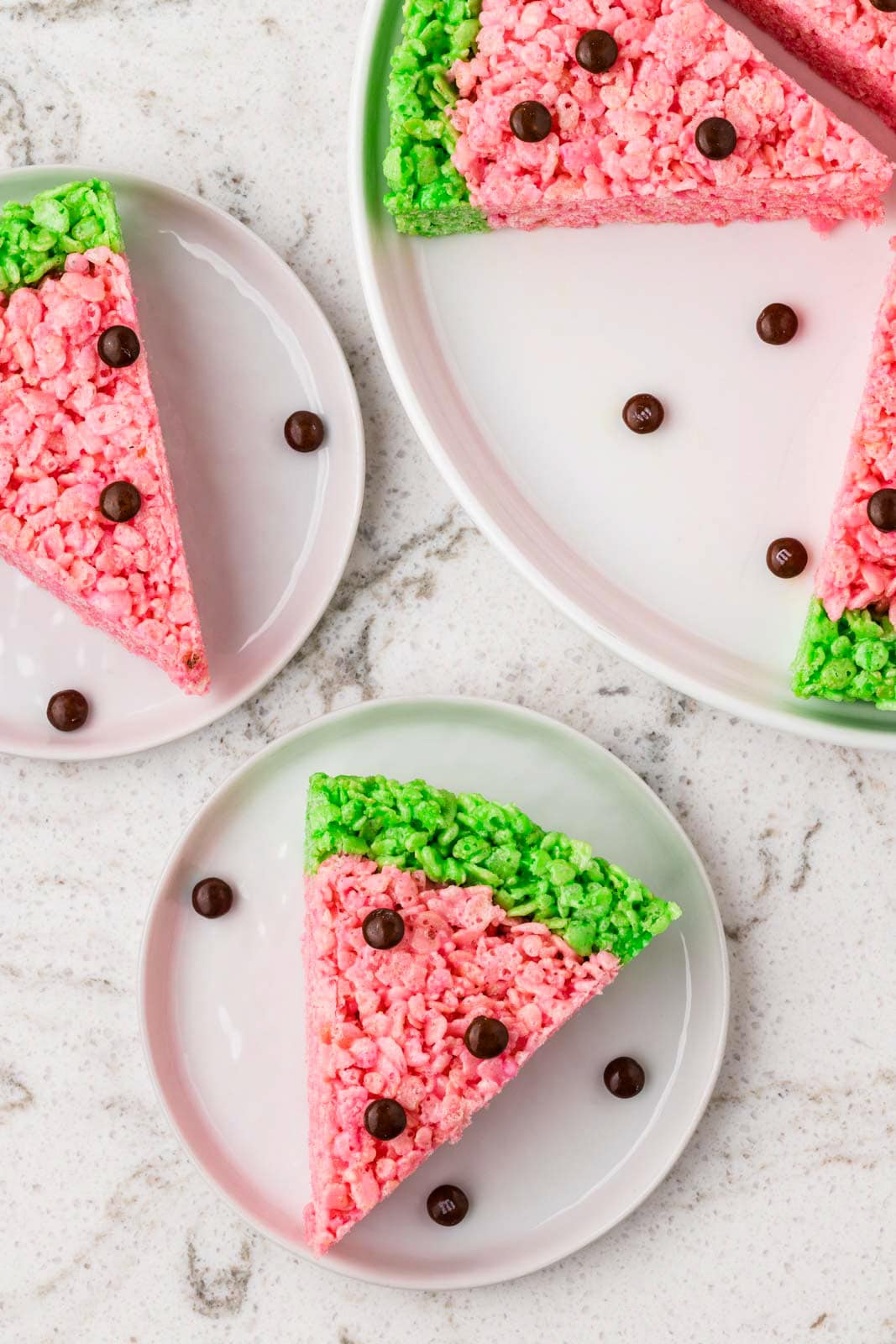 Watermelon-shaped Rice Krispies treat slice on a plate, featuring a pink center, bright green rind, and chocolate candy pieces arranged as watermelon seeds.