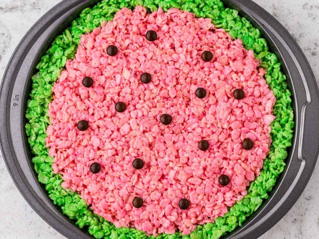 Chocolate candy pieces being placed on top of pink Rice Krispies treats in a round pan to resemble watermelon seeds, with a green rind around the edge.