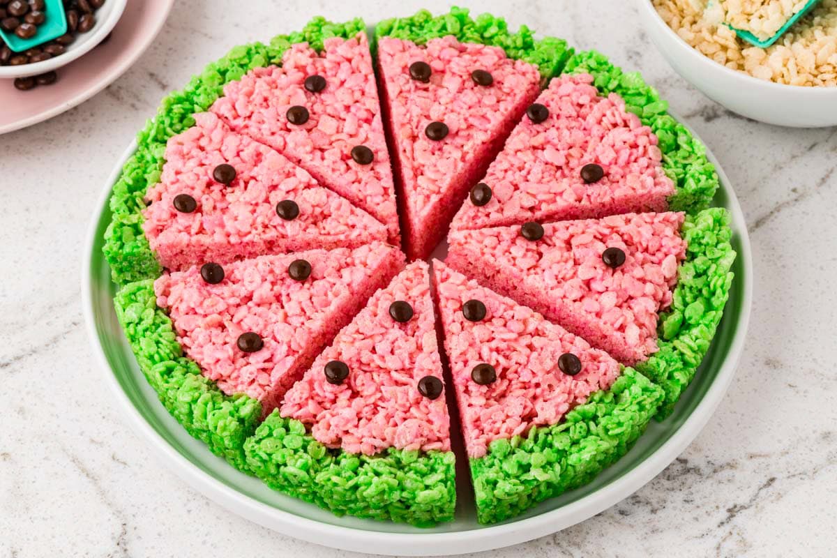 Round watermelon Rice Krispies treat dessert cut into triangular slices and arranged on a serving platter, decorated with a pink center, green rind, and chocolate candy seeds.