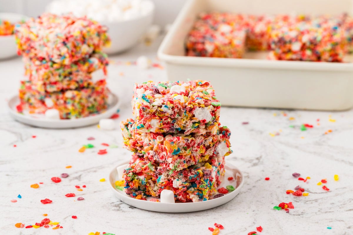 Stack of rainbow Rice Krispies treat squares on a small plate, loaded with colorful cereal pieces, mini marshmallows, and candy-coated chocolates, with more treats in the background.