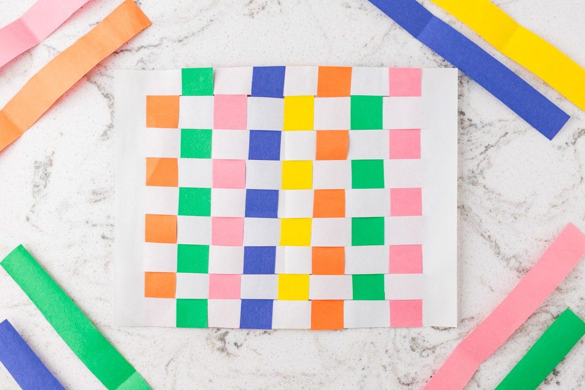 Colorful paper weaving craft made with pastel strips woven through white paper, surrounded by extra paper strips.