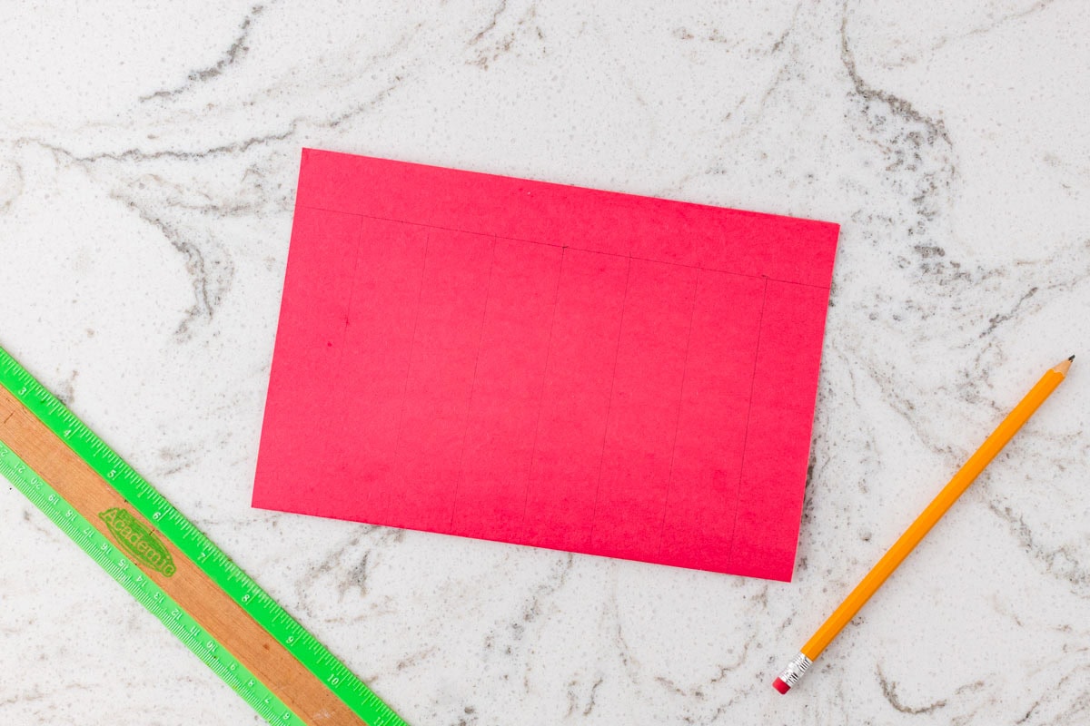Red construction paper marked with evenly spaced vertical cutting lines, a ruler, and a pencil for a paper weaving craft.