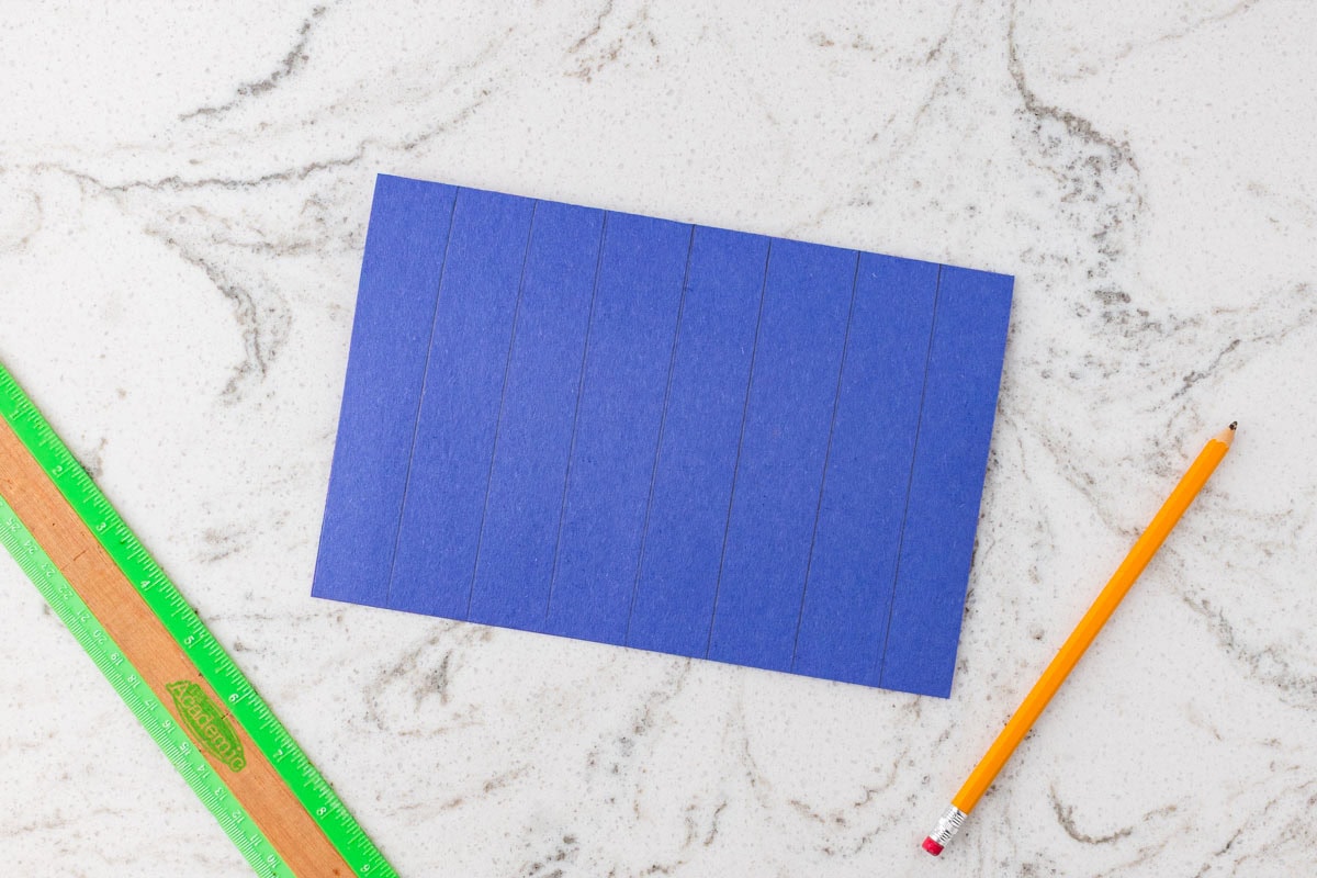 Blue construction paper marked with vertical cutting lines, placed beside a ruler and pencil before cutting.