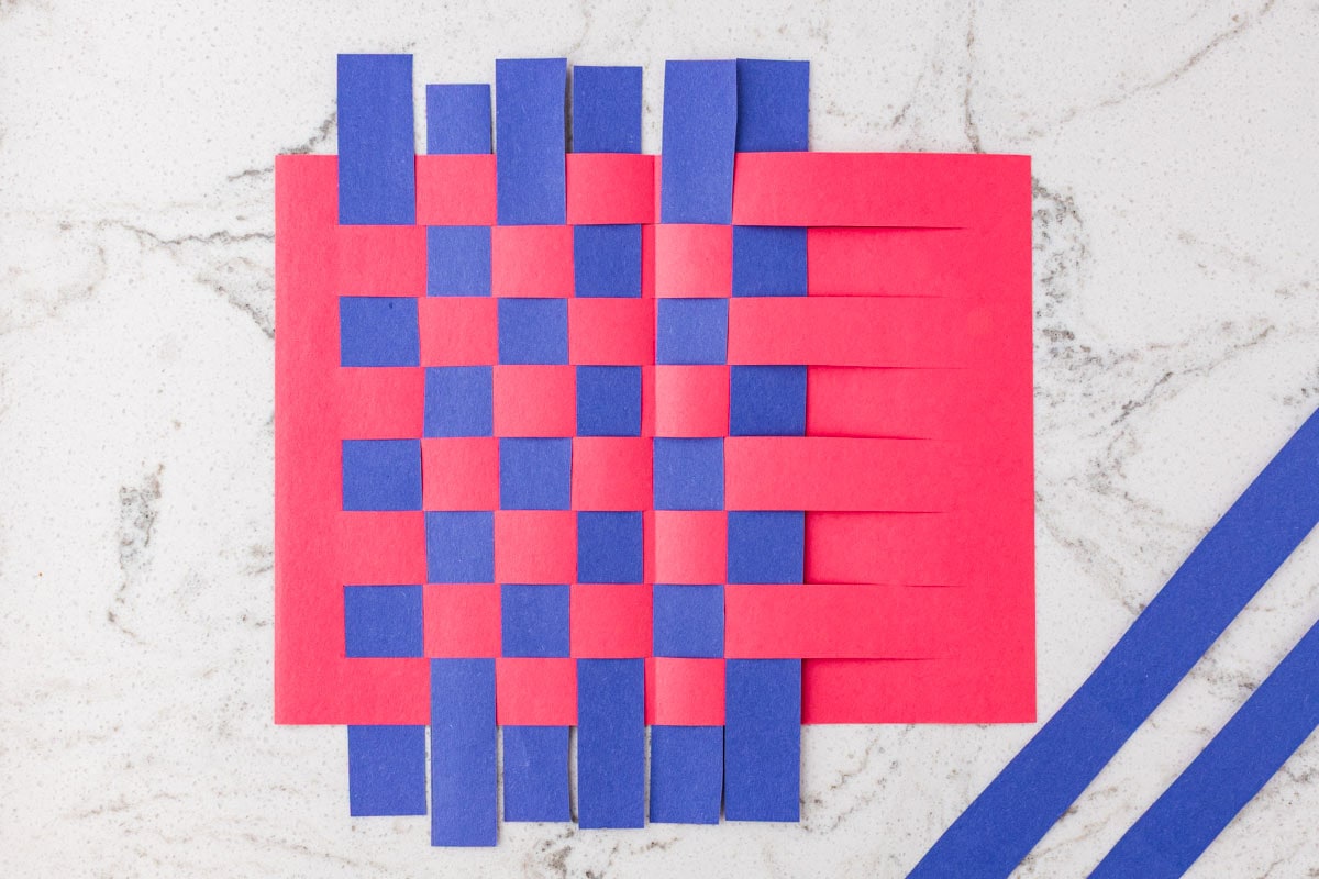 Blue paper strips woven through slits in red construction paper to create a checkerboard paper weaving pattern.
