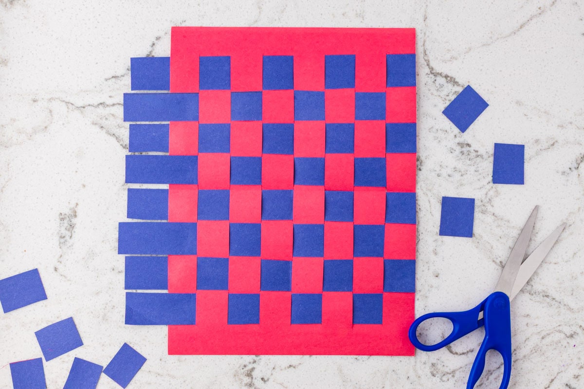 Nearly completed red and blue paper weaving craft with scissors and extra paper squares beside the woven design.