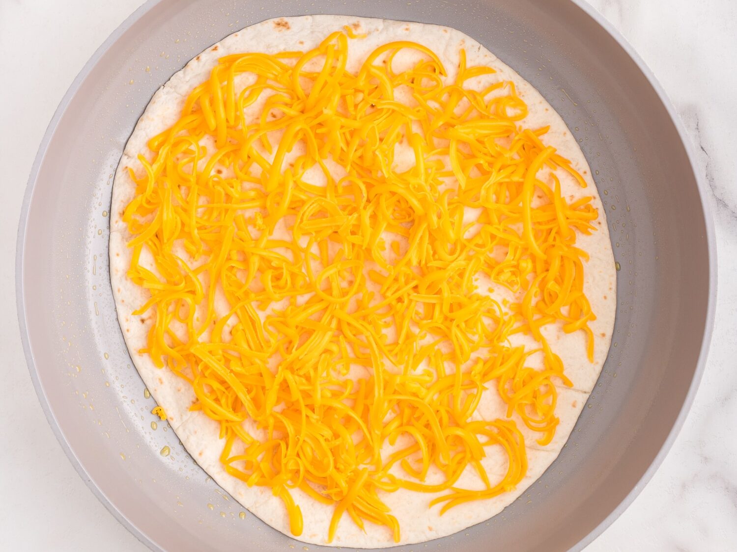 Large tortilla layered with shredded cheese in stovetop skillet for Cheese Quesadilla