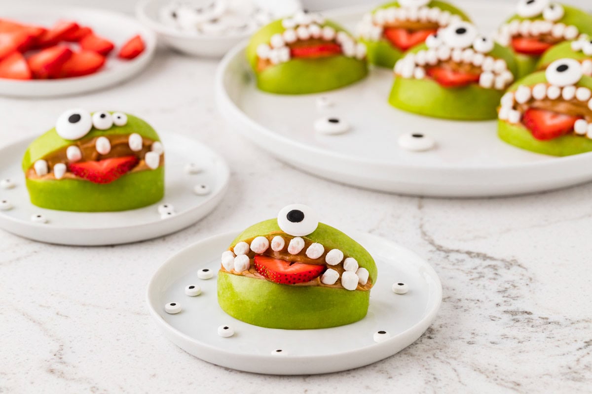 Plates of healthy Halloween apple monster snacks made with green apples, peanut butter, strawberries, mini marshmallows, and edible candy eyes.