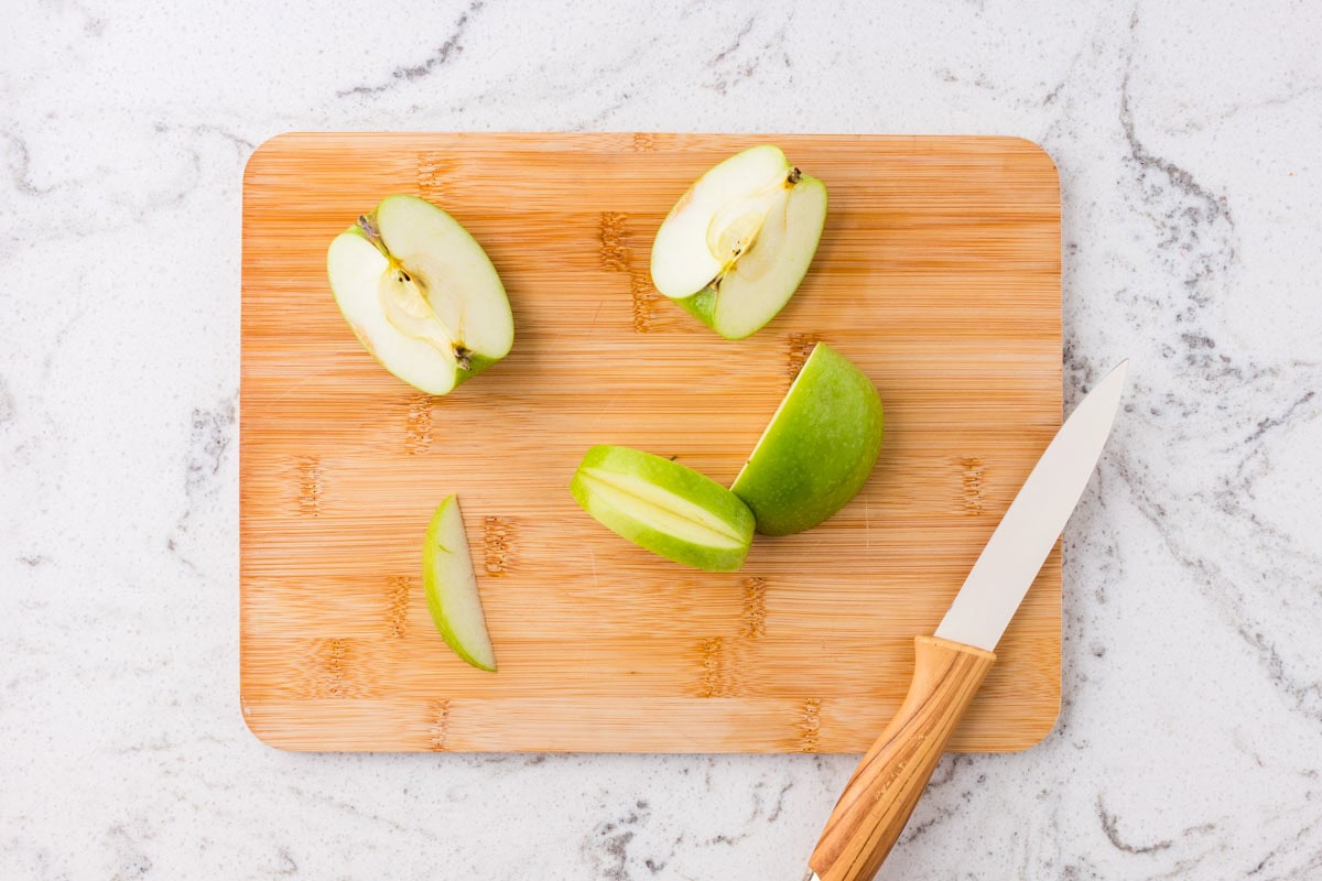 Green apple cut into wedges on a wooden cutting board with a knife, preparing ingredients for Halloween apple monster snacks.