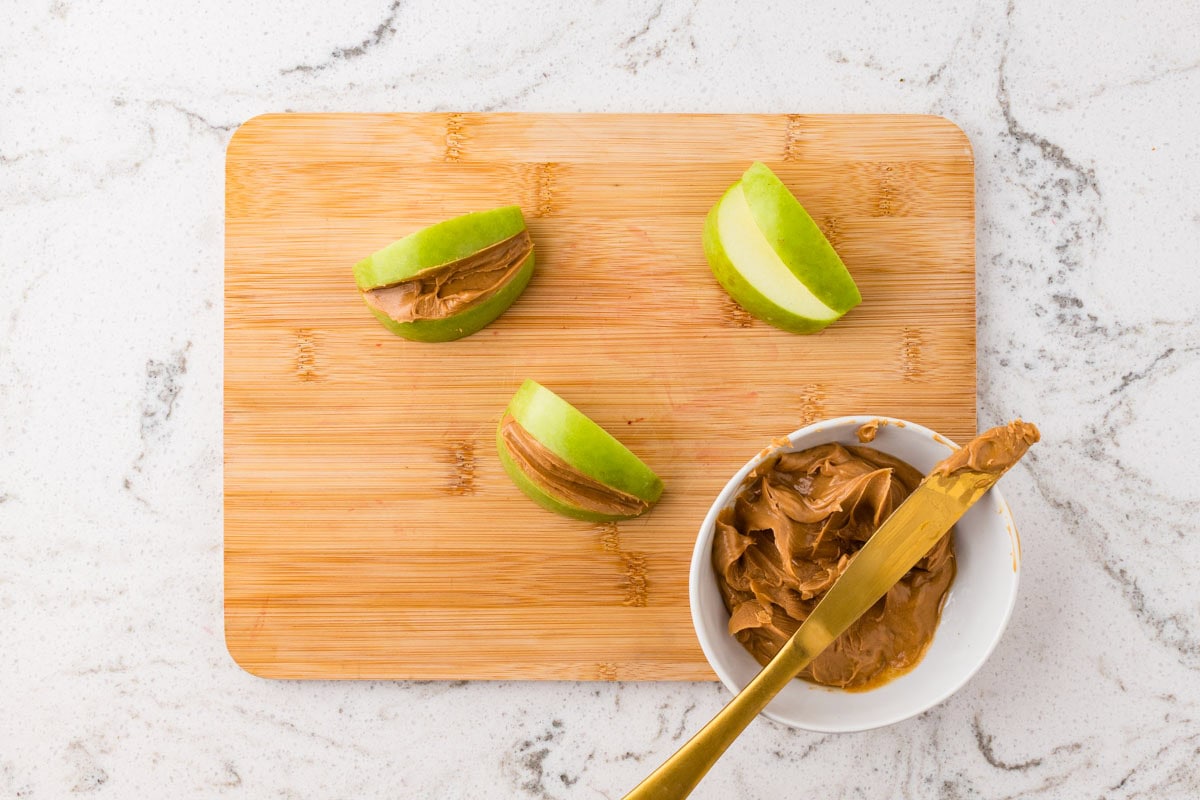 Green apple slices spread with peanut butter on a cutting board beside a bowl of peanut butter for easy Halloween treats.
