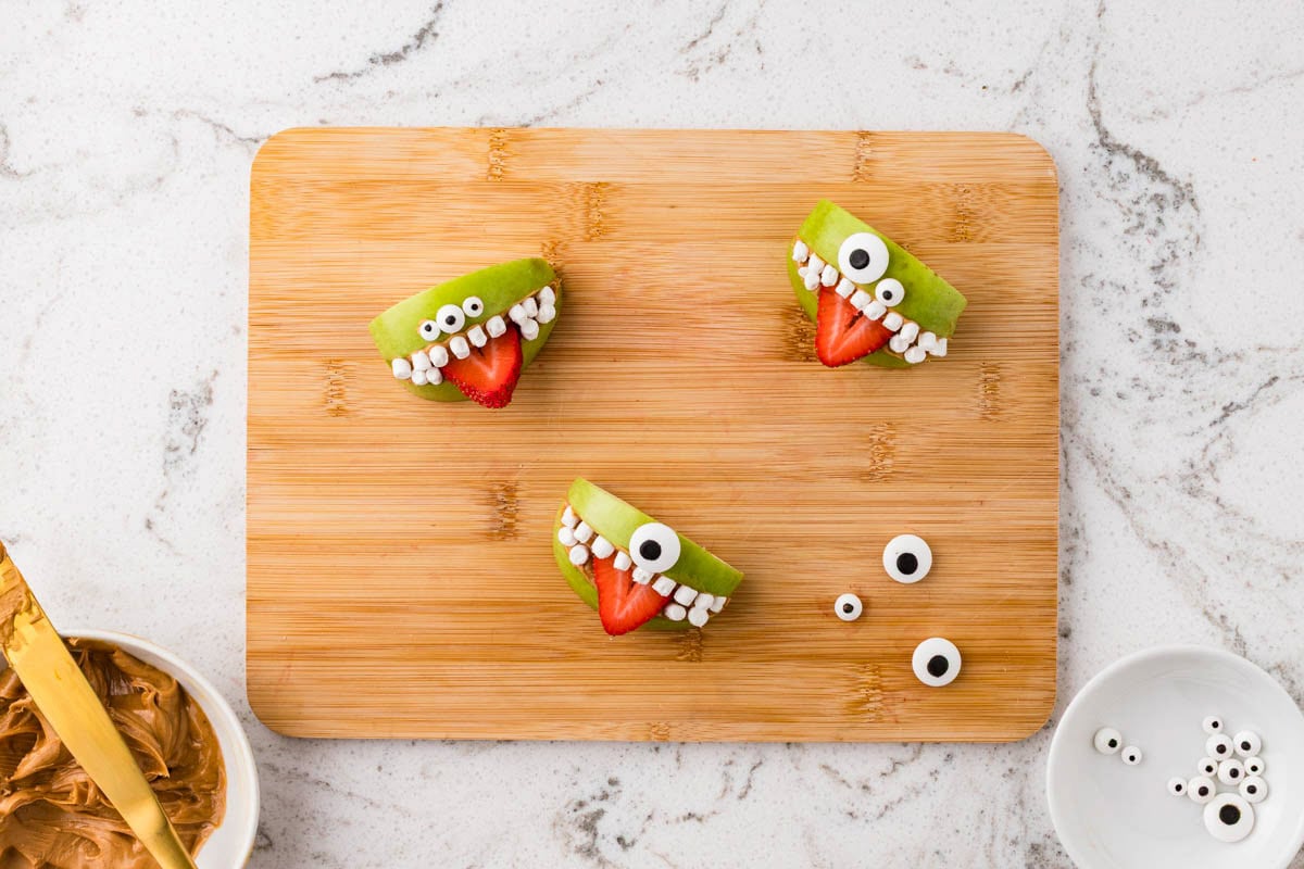 Apple monster snacks decorated with mini marshmallows for teeth and candy eyes on a cutting board for a fun Halloween treat.