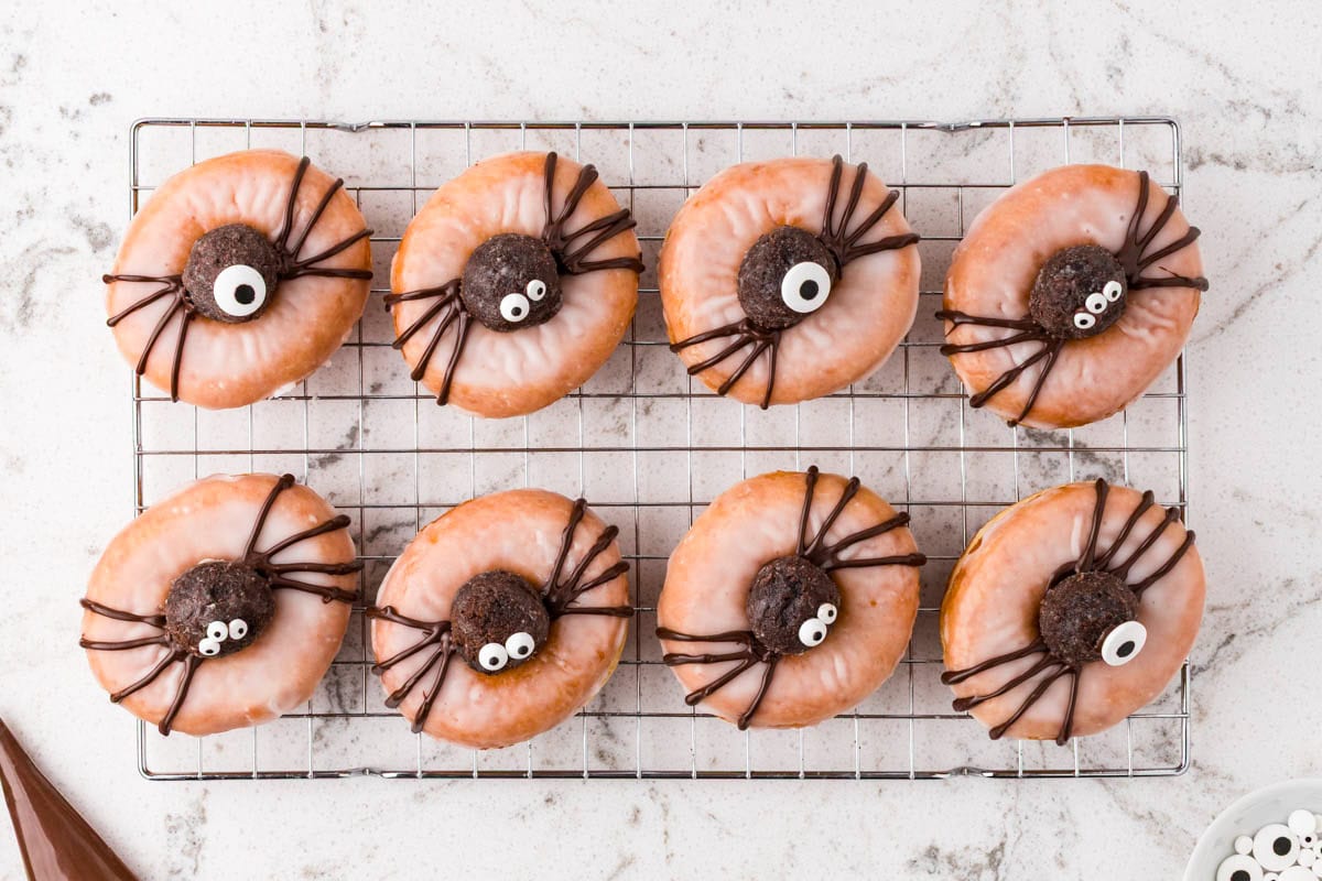 Halloween spider donuts decorated with chocolate legs and assorted candy eyes on a cooling rack.