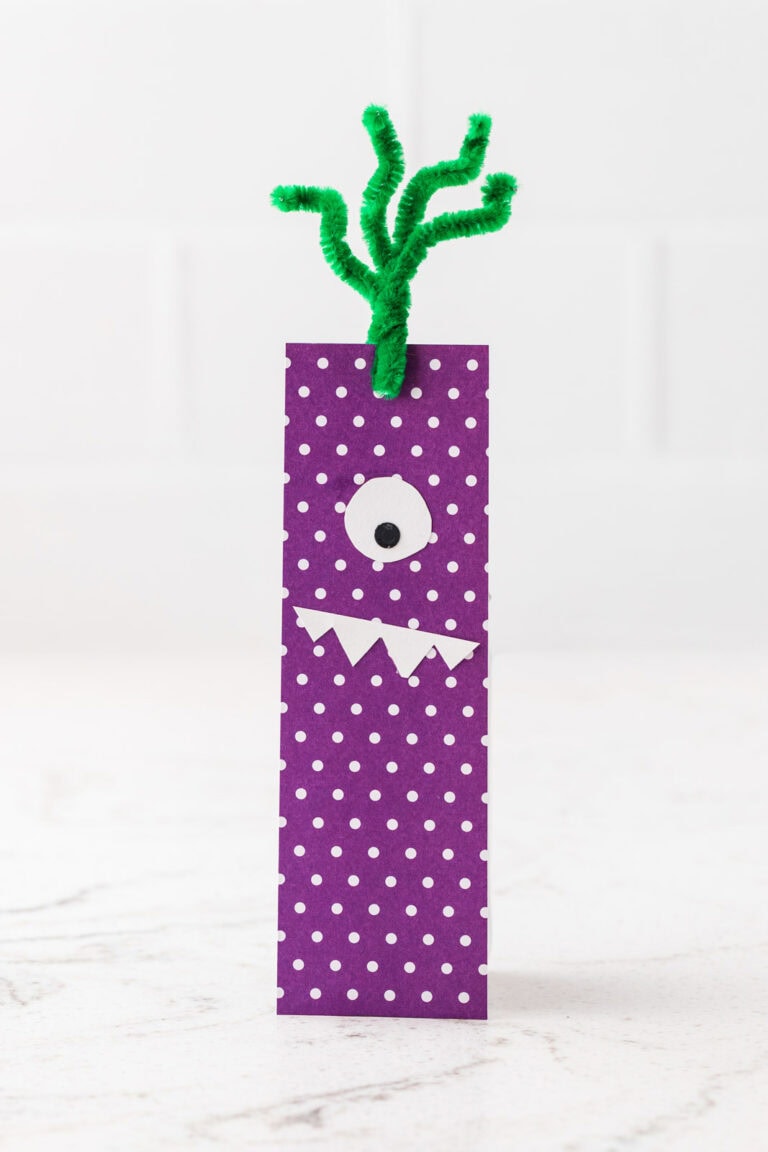 Purple polka-dot monster bookmark with one eye, white pointed teeth, and green pipe cleaner tentacles.