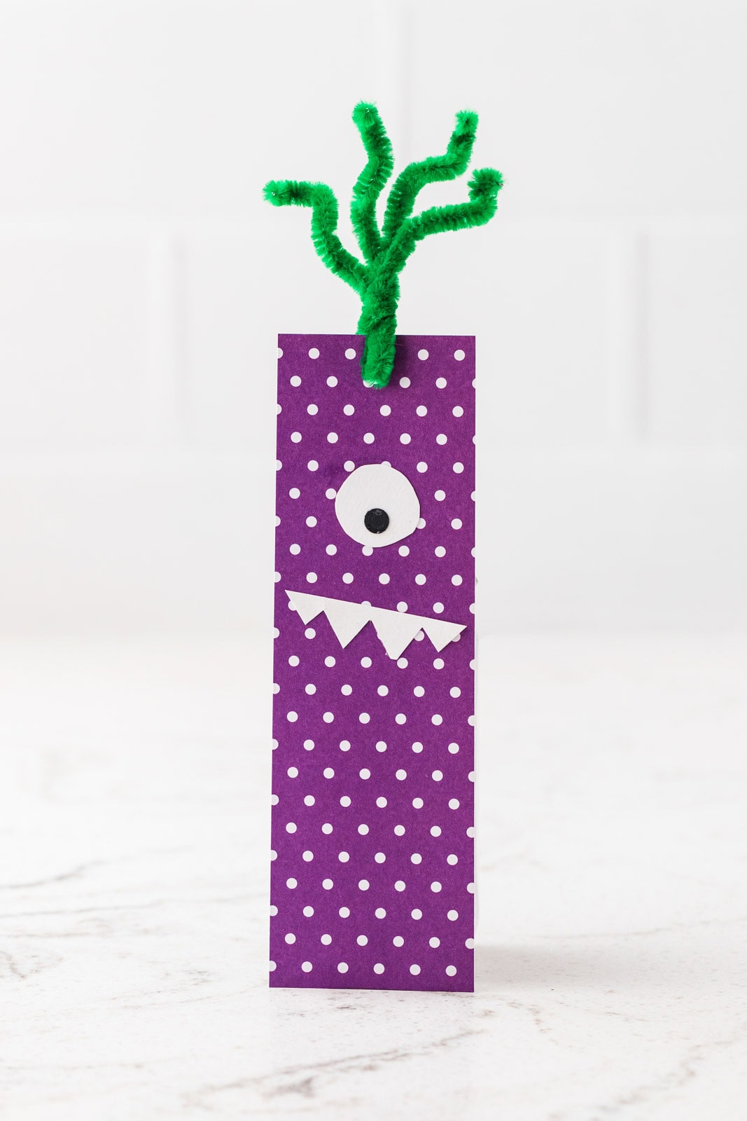 Purple polka-dot monster bookmark with one eye, white pointed teeth, and green pipe cleaner tentacles.
