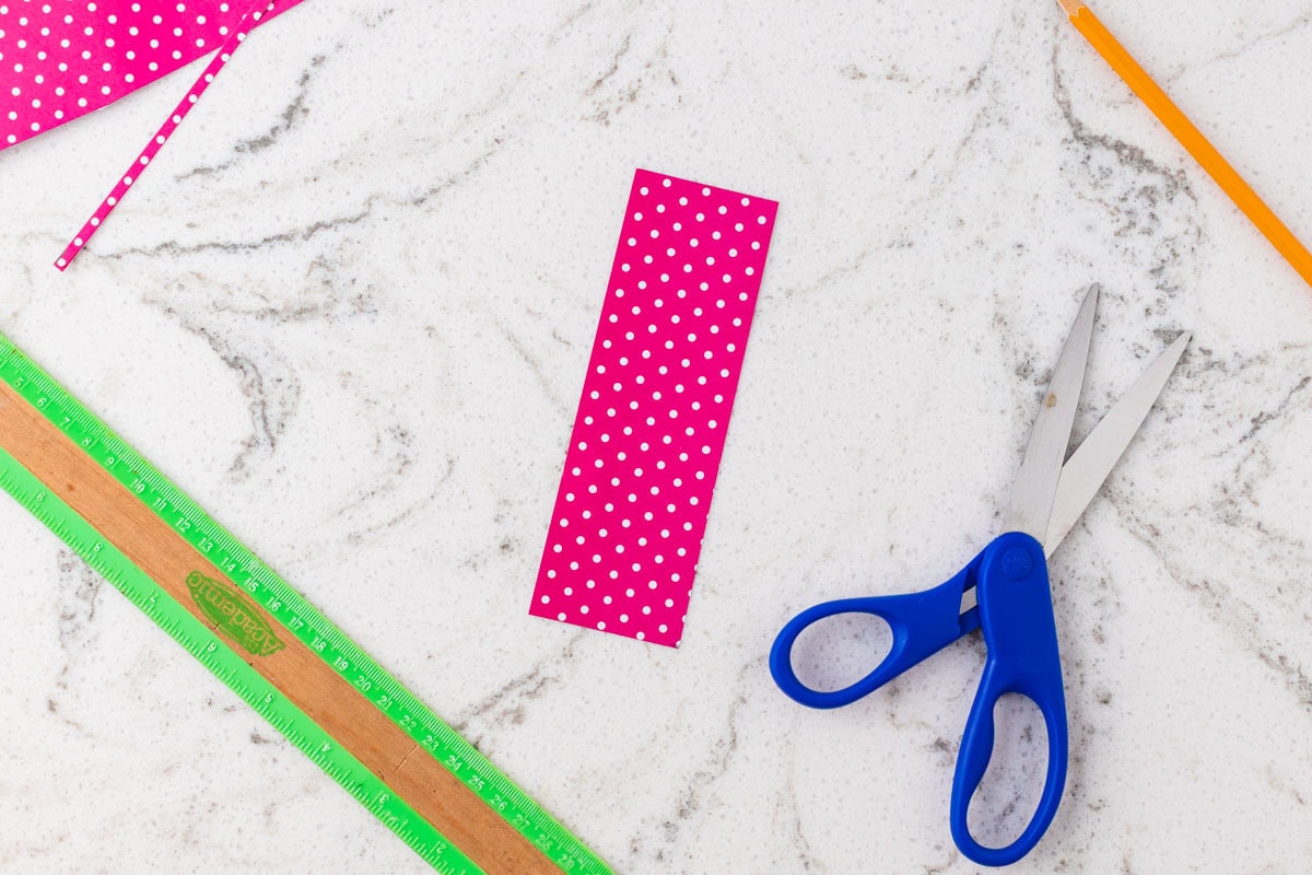 Pink polka-dot paper strip cut into a bookmark, with scissors, ruler, and pencil nearby.