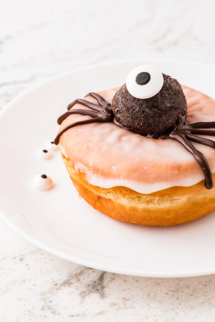 Close-up of a glazed Halloween donut topped with a chocolate truffle spider, a large candy eye, and piped chocolate legs.