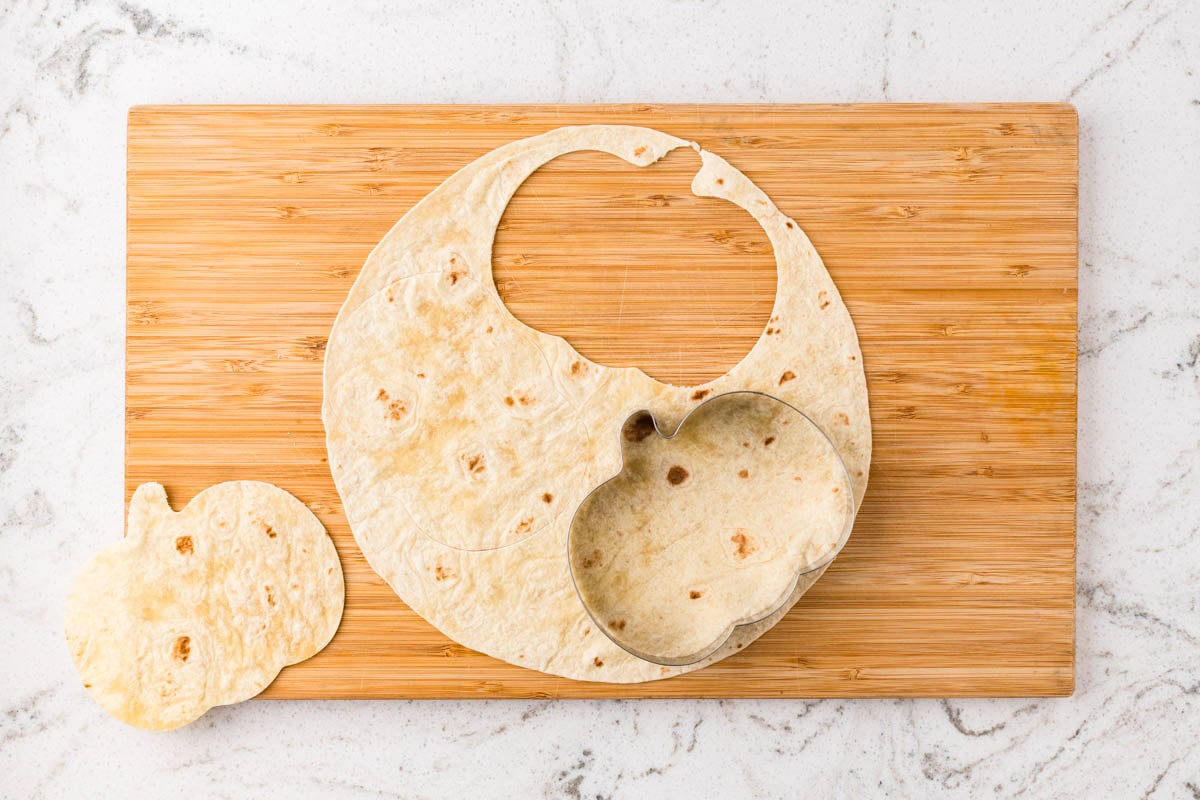 Flour tortilla cut into a pumpkin shape using a pumpkin cookie cutter on a wooden cutting board.