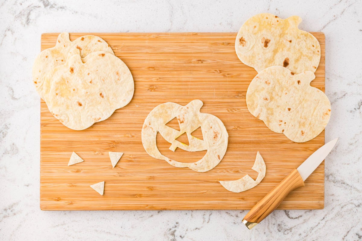 Pumpkin-shaped tortillas with triangle eyes and smiling jack-o’-lantern faces being cut out with a knife.