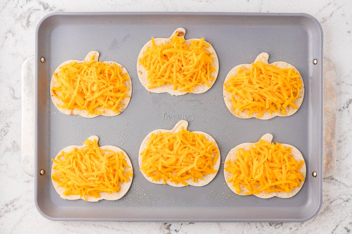 Pumpkin-shaped tortillas topped with shredded cheddar cheese on a baking sheet.