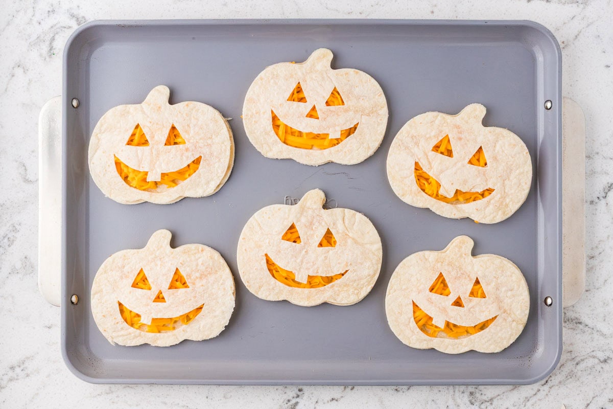 Jack-o’-lantern tortilla cutouts placed over cheese-filled pumpkin-shaped tortillas on a baking sheet.