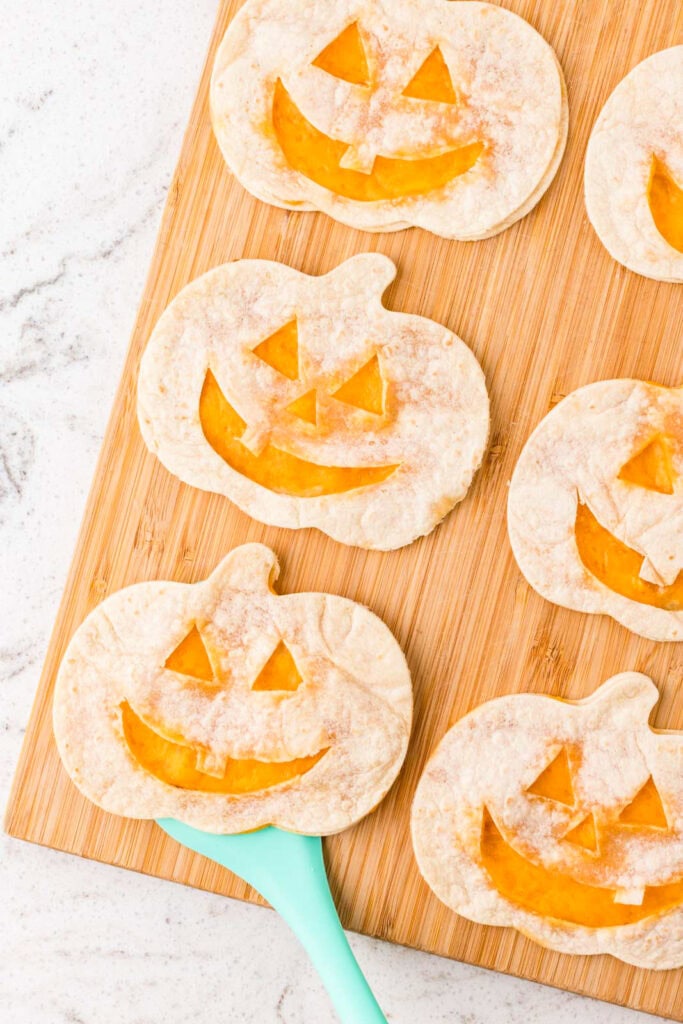 Pumpkin-shaped quesadillas with smiling jack-o’-lantern faces and melted cheddar cheese being lifted from a wooden cutting board.